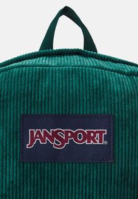 Green corduroy backpack featuring a navy label with "JANSPORT" in white text. Rounded top and fabric texture are prominent.