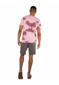 T-shirt rosa a maniche corte in cotone con motivo palme rosa scuro; abbinato a pantaloncini di jeans grigi; modella di spalle, che indossa sandali marroni.