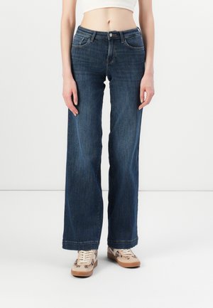 Weite Jeans aus dunkelblauem Denim mit glatter Textur, fünf Taschen und einem klassischen Knopf- und Reißverschluss. Kombiniert mit gemusterten Schuhen.