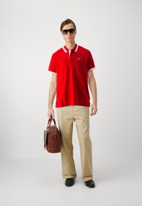 Camisa polo roja con cuello blanco contrastante, pantalones caqui y bolso de cuero marrón. Gafas de sol oscuras y zapatos negros completan el conjunto.