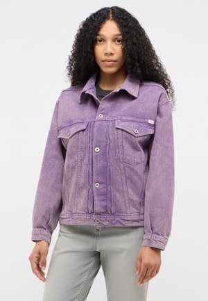 Junge Frau mit lockigem Haar, die eine ausgewaschene lila Jeansjacke und hellgraue Jeans trägt, steht vor einem einfarbigen Hintergrund.