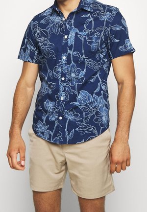 Homme portant une chemise bleu marine à manches courtes avec imprimé floral blanc et un short beige jusqu'aux genoux, debout devant un fond blanc.