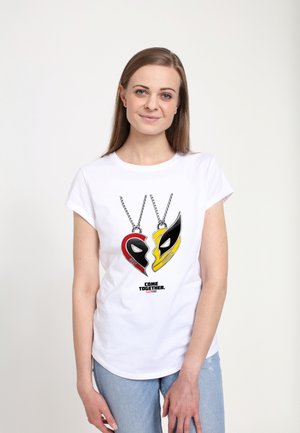 Ung kvinde iført en hvid t-shirt med to superheltemaske-vedhæng og ordene "Come Together" på en ensfarvet baggrund.