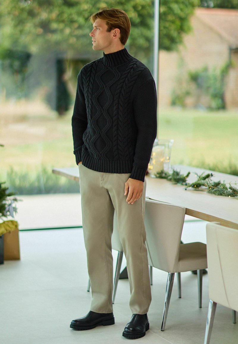 Zwarte kabelgebreide turtleneck trui met ribbelrand, gecombineerd met beige broek en zwarte enkellaarsjes, geplaatst in een moderne binnenomgeving.