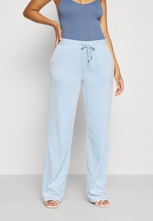 Femme portant un pantalon large bleu clair avec une taille à cordon et des sandales blanches ouvertes aux orteils, associées à un haut bleu sans manches.