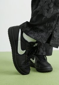 Sapatilhas de couro preto com logo Swoosh prateado, painéis texturizados e sola de borracha preta. Usadas com calças pretas estampadas, repousando sobre uma superfície verde.
