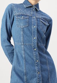 Abito camicia in denim azzurro chiaro con silhouette aderente, caratterizzato da due tasche al petto e dettagli di borchie trasparenti sulle spalle.
