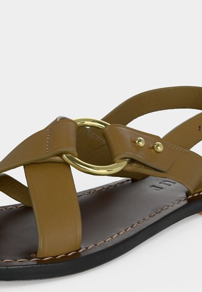 Brūnas ādas sandales ar zelta apaļu sprādzi, melnu zoli un redzamām šuvēm gar malu.