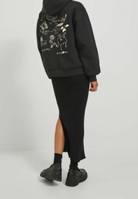 Svart hoodie med gyllene grafiska mönster på baksidan, kombinerad med en svart ribbad nederdel med en sidoklyvning. Kraftiga svarta skor fulländar looken.