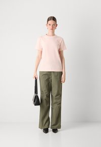 T-shirt di cotone rosa chiaro con maniche corte. Pantaloni oliva a vestibilità ampia con una leggera lucentezza. Borsa a mano nera strutturata e scarpe nere a punta.