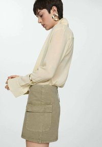 Khaki denim rok met zijzakken, gecombineerd met een losse, crèmekleurige zijden blouse met brede manchetten. Gouden oorbellen met een ring accentueren de look.