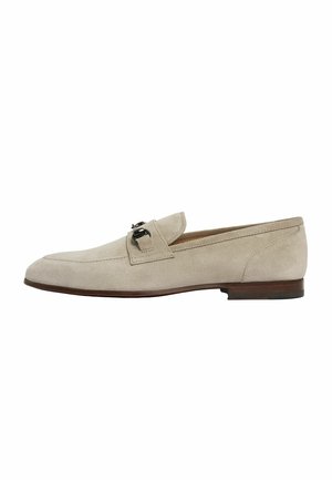 Mocasin de ante beige con una textura suave, puntera redondeada y detalle decorativo de hebilla en la parte delantera. Tacón plano y diseño minimalista.
