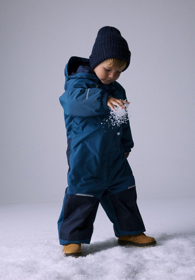 Niño pequeño con un traje de nieve azul y un gorro de punto azul marino jugando con nieve artificial en un suelo blanco con fondo claro.