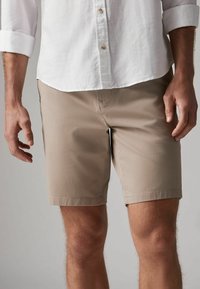 Shorts beiges en tissu léger, présentant une coupe droite, un devant plat et un ourlet piqué. Portés avec une chemise blanche à boutons.