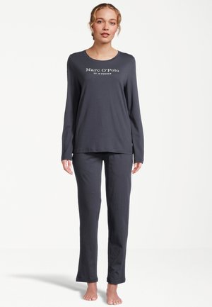 Vrouw staat blootsvoets, draagt een donkergrijze top met lange mouwen en bijpassende broek met de tekst "Marc O'Polo" op de borst tegen een witte achtergrond.