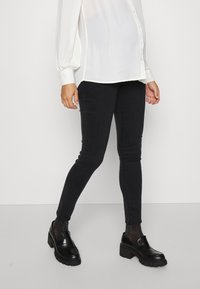 Jeans skinny pretos em tecido elástico, combinados com uma blusa branca de corte solto. Footwear apresenta mocassins pretos brilhantes com solas grossas.