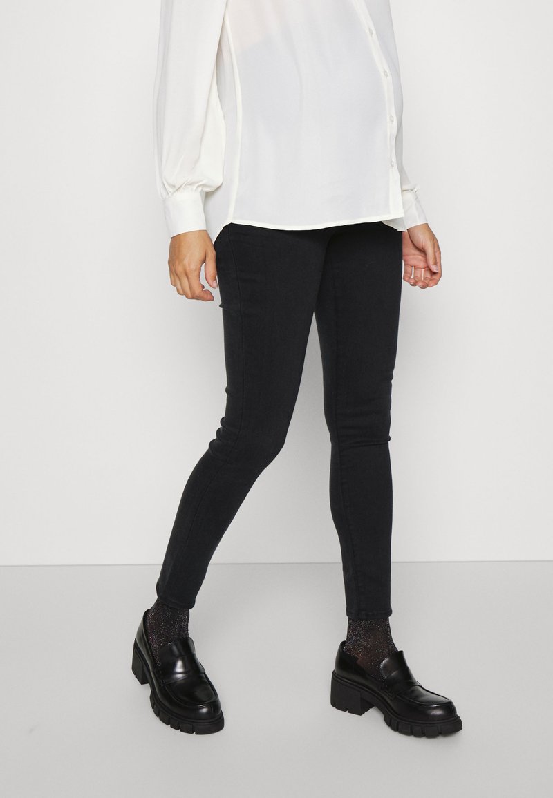 Jeans skinny pretos em tecido elástico, combinados com uma blusa branca de corte solto. Footwear apresenta mocassins pretos brilhantes com solas grossas.