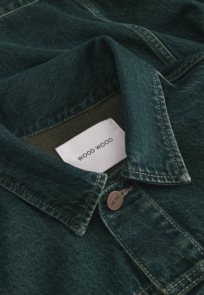 Kołnierz ciemnozielonej kurtki dżinsowej z widoczną białą metką "WOOD WOOD" oraz metalowym guzikiem z wygrawerowanym napisem "W.W."