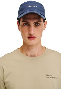 Niebieska czapka z napisem "HALO" z przodu, noszona przez mężczyznę. Na sobie ma beżowy t-shirt z czarną naszywką po lewej stronie.