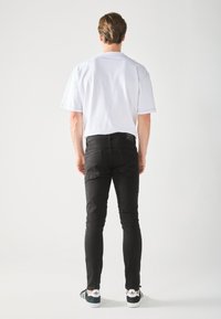 Muži stojaci chrbtom, oblečený v bielej košeli s krátkym rukávom, čiernych džínsoch a čiernych teniskách Adidas s bielymi podrážkami.