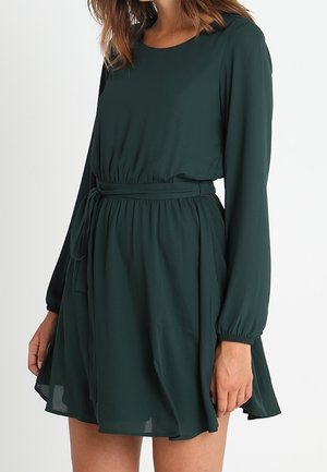 Vrouw die een donkergroene jurk met lange mouwen draagt, met een ronde halslijn en een tailleceintuur, staand tegen een effen witte achtergrond.