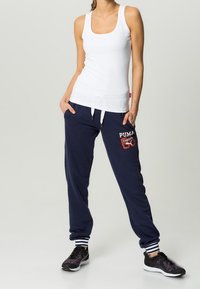 White sleeveless tank top, navy sweatpants with white stripes and red Puma logo, black athletic shoes with purple accents.