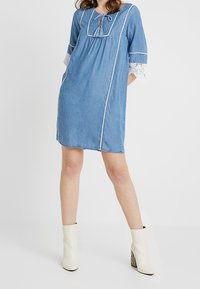 Džínová šaty s přesným výstřihem, krajkovými akcenty na rukávech a stahovacími detaily vpředu. Barva je světle modrá s bílým prošitím.
