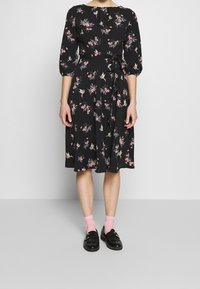 Svart klänning med trekvartslånga ärmar, blommigt mönster i rosa och grönt, midjeband, knälång kjol; bärs med rosa strumpor och svarta skor.