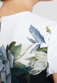 T-shirt à motif floral avec un fond blanc, des feuilles bleues et des fleurs grises. Le design comprend une fermeture éclair dorée au dos.
