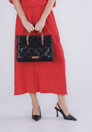Donna in abito midi rosso che tiene una borsa nera in pizzo floreale con manici marroni, indossando décolleté slingback nere con punta appuntita.