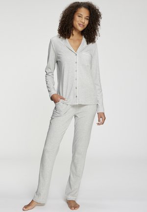 Ženska stoji bosa, oblečena v svetlo sivo pižamo z dolgimi rokavi in gumbi ter ujemajoče pižamke hlače z žepi.