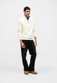 Cream-colored half-zip sweatshirt with a navy accent at the collar, paired with black straight-leg jeans and tan shoes. Smooth texture and casual fit.