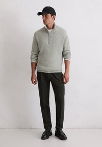 Pull en maille côtelée gris clair avec un col zippé, associé à un pantalon vert foncé et des mocassins noirs, sur un fond neutre.
