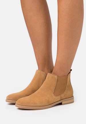 Jambes portant des bottines Chelsea en daim beige avec des panneaux latéraux élastiques et des talons bas empilés sur un fond blanc.