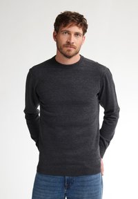 Pulover gri închis, cu guler rotund, fabricat dintr-un material moale, cu o textură netedă. Mâneci lungi și manșete cu bandă, purtat cu blugi casual albaștri.