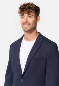 Sötétkék zakó, puha anyagból, slim fit szabással, egy gombos záródással, kihajtott gallérral és zsebrevalóval. Fehér póló fölött viselve.