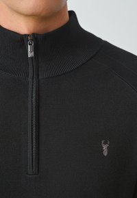 Maglione zip-up nero con colletto alto, caratterizzato da un motivo a maglia testurizzata e un piccolo logo di cervo sul petto. La cerniera è di tonalità argentata.