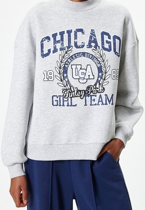 Sweat-shirt gris avec texte bleu indiquant "Chicago Girl Team," "USA," "Athletic Division," et "Tinley Park", porté avec un pantalon bleu.