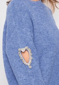 Pull en tricot bleu avec un col rond, doté d'une découpe en forme de cœur unique sur la manche bordée de perles décoratives.