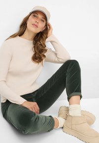 Beige ribstrikket sweater, mørkegrønne fløjlsbukser, beige slip-on støvler og en lysebeige kasket med blå accent. Person der sidder afslappet.