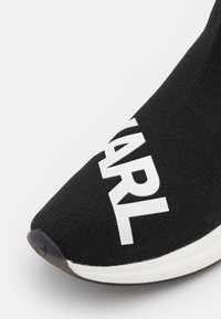 Svart sneaker med ett elastiskt ovandel av tyg, med stora vita bokstäver och en slät gummisula. Rund tå och minimalistisk design.