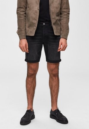 Mężczyzna w czarnych dżinsowych szortach podwiniętych na mankietach, brązowej zamszowej kurtce, czarnej koszuli i czarnych sznurowanych butach, stojący na jednolitym tle.