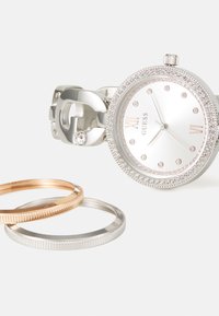 Orologio argento con quadrante rotondo, dettagli in cristallo e numeri romani. Include due anelli: uno argento e uno oro rosa, con dettagli texture.