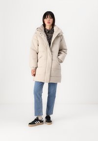 Manteau doudoune beige avec un col haut, des poches zippées et des poignets élastiques ; porté sur une chemise à motifs foncés, associé à un jean bleu et des baskets noires.