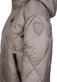 Giacca a vento beige con texture trapuntata, tasca laterale con zip e una toppa rotonda con logo sulla spalla sinistra. Design con cappuccio.
