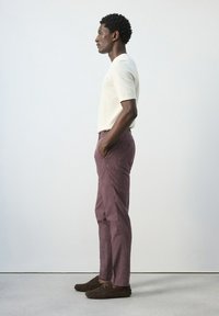 Hombre de perfil con las manos en los bolsillos, vestido con una camisa blanca de manga corta, pantalones morados a rayas y mocasines marrones, sobre un fondo liso.