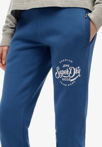 Niebieskie bawełniane dresy z białym nadrukowanym logo na lewym udzie, elastycznym pasem i bocznymi kieszeniami.