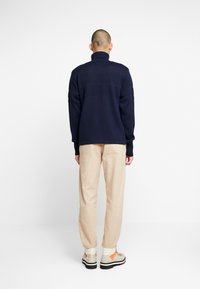 Maglione lavorato a maglia blu navy con colletto alto, righe testurizzate e maniche lunghe, abbinato a pantaloni beige e calzature multicolori.