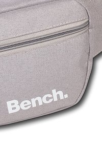 Grijze stoffen tas met een ritssluiting, met een gladde textuur en een wit "Bench." logo dat prominent op de voorkant staat.