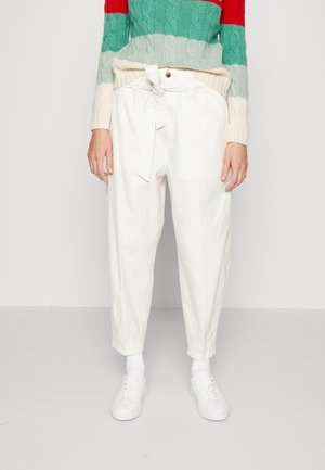 Personne portant un pantalon blanc noué à la taille, des baskets blanches et un pull avec des blocs de couleur rouge, vert et crème, debout devant un fond uni.